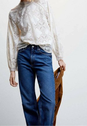 Personne portant un chemisier en dentelle blanche et un jean bleu tenant une veste en daim marron sur fond uni.