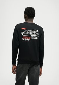 Μαύρη μπλούζα με βαμβακερή σύνθεση, με έντονα κόκκινα και λευκά γραφικά στο πίσω μέρος, συμπεριλαμβανομένων των "REPLAY RACINGTEAM" και "FOR SUPERIOR PERFORMANCE."