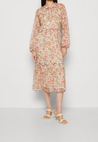 Robe midi fleurie à manches longues, tissu léger, motifs roses et verts, ourlet à volants, et détail noué au col, portée avec des talons beiges.