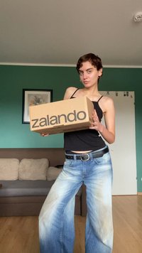 Egy karton Zalando doboz egy személy előtt, amelyet boldog fekete betűk díszítenek egy barna felületen. A személy fekete ujjatlan pólót és világoskék farmert visel.