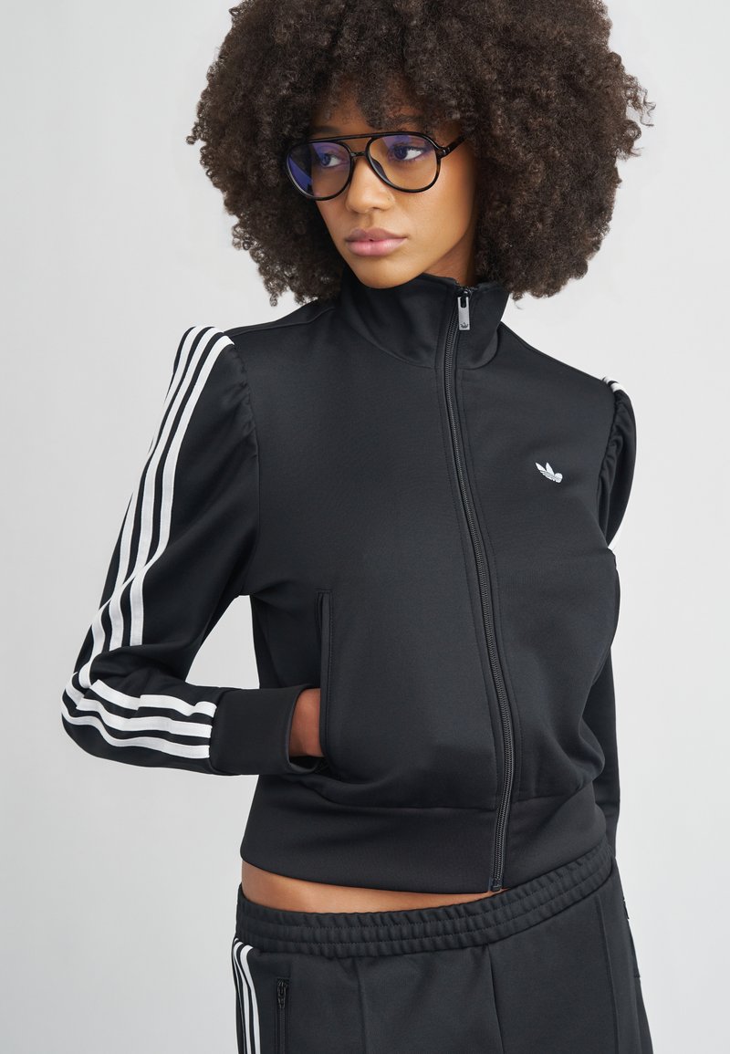 Μαύρο μπουφάν με φερμουάρ και λευκές τρεις ρίγες στα μανίκια. Διαθέτει ψηλό γιακά και πλαϊνές τσέπες, κατασκευασμένο από λείο ύφασμα.