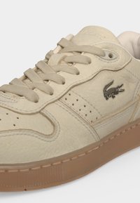 Baskets en cuir beige avec lacets beiges, détails perforés, semelle marron et logo crocodile Lacoste noir sur le côté.