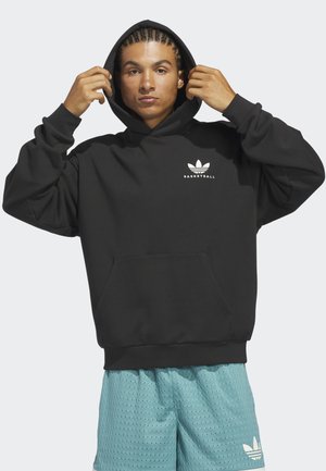 Young man adjusting black hoodie with white basketball logo, wearing teal sports shorts with white Adidas logo on plain background.