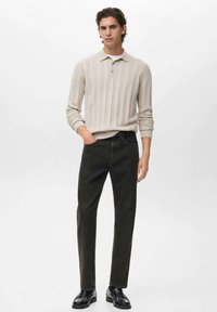 Camisa de polo de rayas beige con cuello, combinada con jeans rectos verdes oscuros y zapatos negros, de pie sobre un fondo neutral.