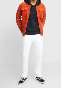 Chaqueta de pana naranja sobre una sudadera negra, jeans blancos y zapatillas Vans negras. Las características destacadas incluyen bolsillos y variaciones en la textura.