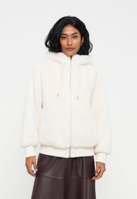 Donna con capelli scuri che indossa una felpa bianca con zip e una gonna in pelle marrone scuro, in piedi davanti a uno sfondo bianco semplice.