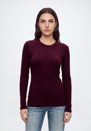 Vrouw met lang donker haar, die een aansluitend bordeauxrood ribgebreid shirt met lange mouwen en lichtblauwe spijkerbroek draagt, staand tegen een effen witte achtergrond.