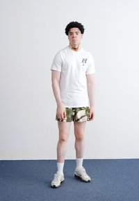 Witte T-shirt met korte mouwen met een "N" logo, groene camouflage shorts, witte enkelkousen en grijze sneakers. Effen achtergrond met blauwe vloer.
