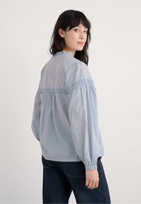 Frau mit dunklem Haar, die über die Schulter schaut, trägt eine hellblaue Bluse mit langen Ärmeln und gesticktem Einsatz sowie dunkle Jeans.