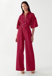 Raudonas struktūruotas šokinėjimas su smailiu apykaklę, priekiniais mygtukais ir pūstomis rankovėmis. Su diržu juosmens ir plačiomis kelnėmis. Lygus paviršius.