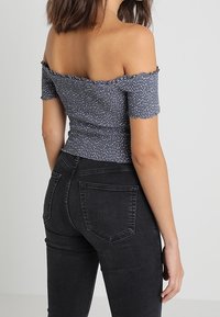 Top sin mangas en azul oscuro con motas blancas, tejido texturizado y bordes con volante; combinado con jeans negros ajustados que tienen bolsillos traseros.