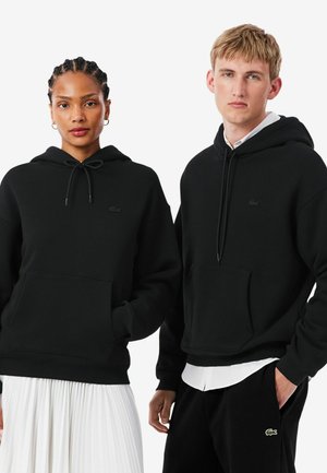 Deux personnes portent des sweats à capuche noirs, l'une associée à une jupe plissée blanche et l'autre à un pantalon noir avec une chemise blanche en dessous.