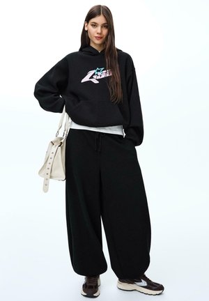 Giovane donna in felpa nera oversize con grafica, pantaloni neri larghi, sneakers marroni, che porta una borsa beige su sfondo bianco.