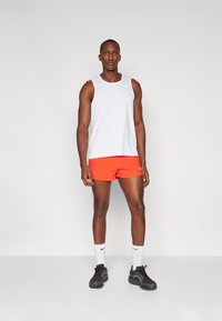 Herreidrettsklær med en hvit ermeløs topp og oransje shorts, med et teksturert mønster. Svarte joggesko og hvite sokker med logoer synlige.