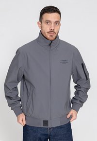 Graue Bomberjacke mit hohem Kragen, Reißverschlusstaschen und elastischen Bündchen. Merkmale ein Logo auf der Brust und eine feste Stofftextur.