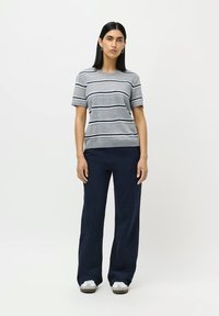 Woman standing wearing a grey short-sleeve striped shirt, navy wide-leg pants, white sneakers, and silver hoop earrings against a white background.