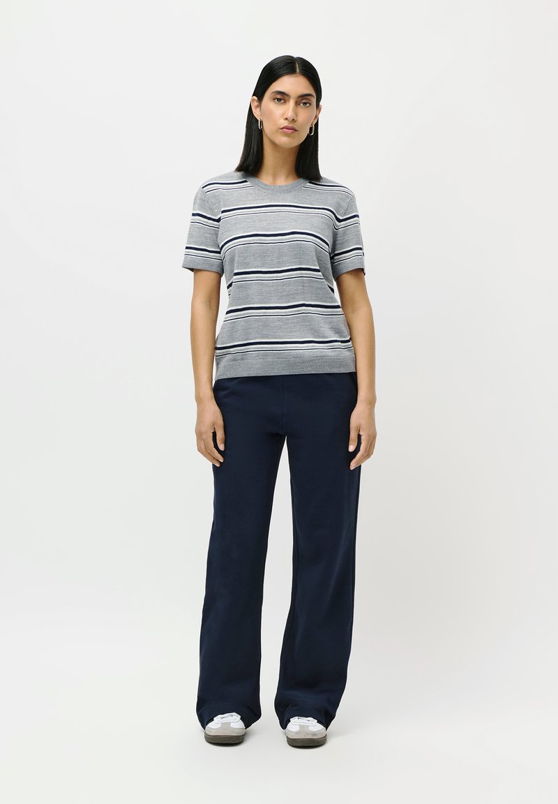 Woman standing wearing a grey short-sleeve striped shirt, navy wide-leg pants, white sneakers, and silver hoop earrings against a white background.