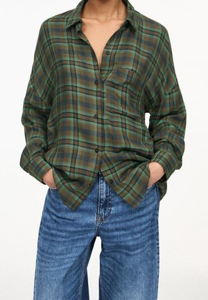 Person wearing a loose green and brown plaid button-up shirt tucked partially into high-waisted blue jeans with hands in pockets.