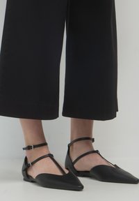 Ballerine nere a punta con doppie cinghie alla caviglia indossate con pantaloni neri a gamba larga e cropped su un pavimento bianco.