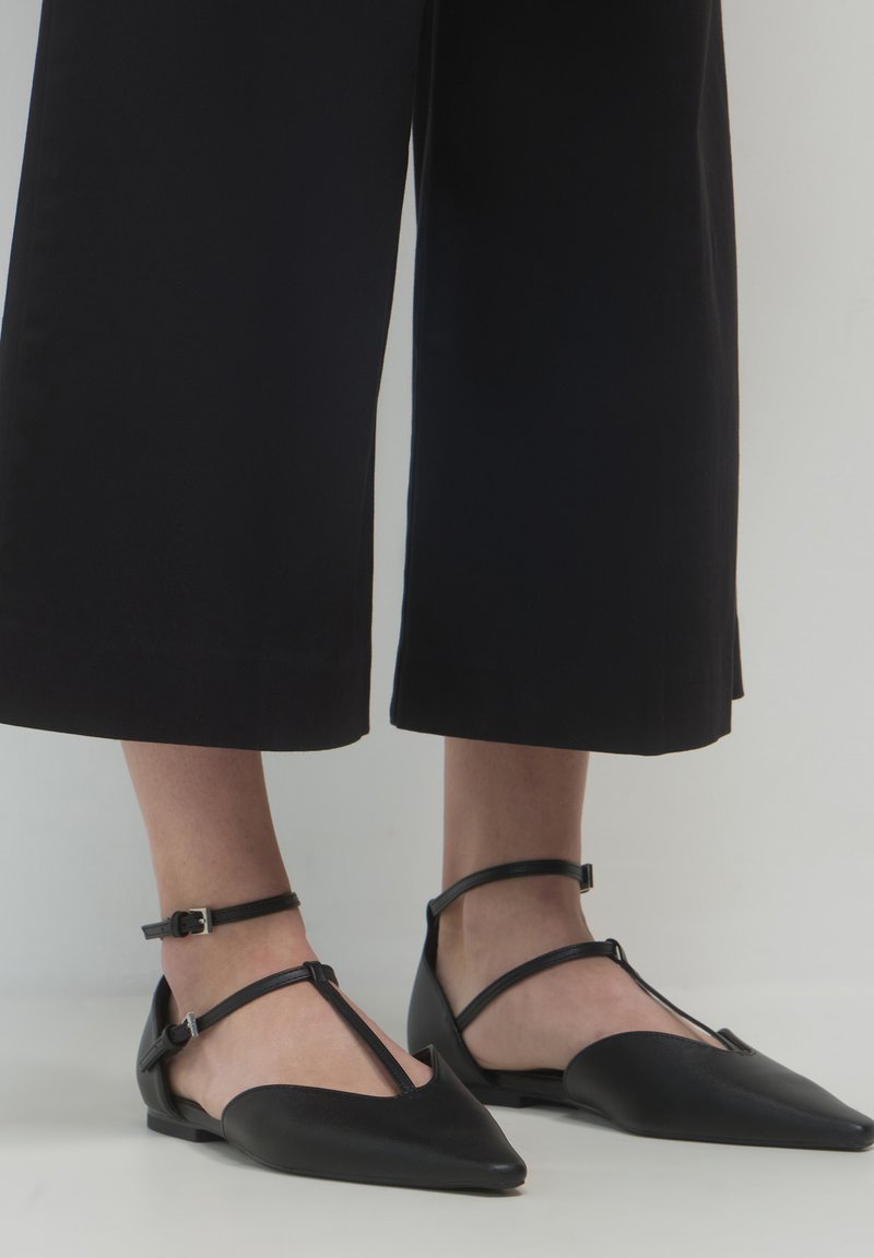 Schwarze spitze Ballerinas mit doppelten Knöchelriemen, getragen mit schwarzen weit geschnittenen cropped Hosen auf einem weißen Boden.