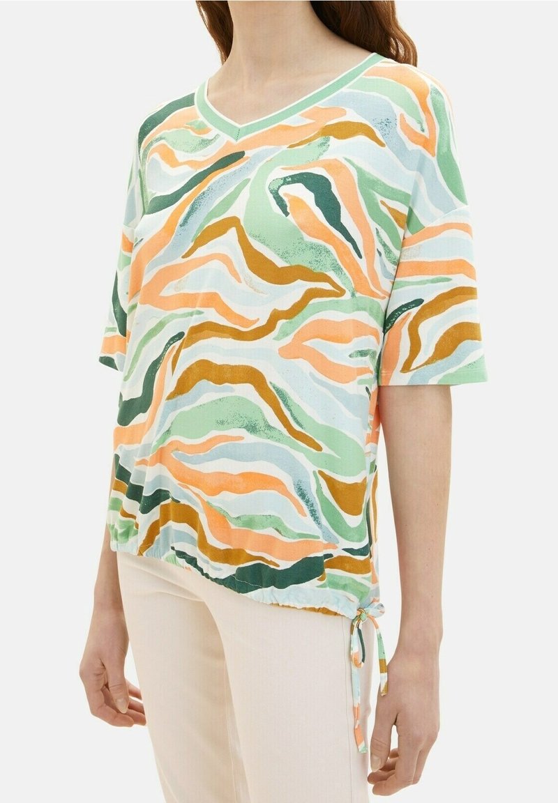 Femme portant un haut à manches courtes et col en V avec des motifs abstraits ondulés verts, orange, beige et bleu clair, noué à l'ourlet sur un côté.