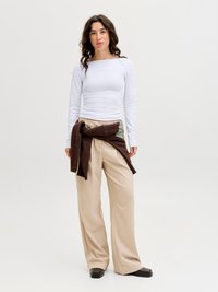 Femeie cu păr creț, purtând un top alb cu mâneci lungi, pantaloni largi bej, pantofi negri și un pulover maro legat în jurul taliei.