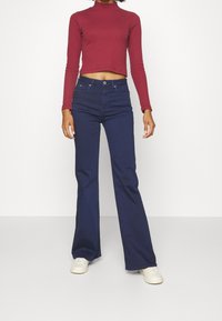 Meri flared jeans dengan pinggang tinggi berwarna navy, dipasangkan dengan atasan lengan panjang cropped warna burgundy. Model mengenakan sepatu sneakers putih, dengan latar belakang polos.
