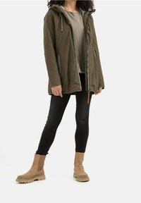 Veste zippée vert olive avec capuche, associée à une chemise grise, un jean noir et des bottines en daim beige. Dotée de détails zippés et de poches.