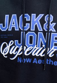 Felpa con cappuccio blu navy con grande testo bianco e blu: "JACK & JONES," "Superior" e "New Aesthetic." Dotata di coulisse con punte metalliche.
