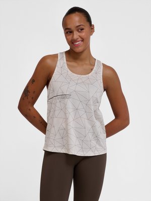 Lächelnde Frau mit Tattoos, die ein weißes ärmelloses Oberteil mit geometrischem Muster und dunkle Leggings trägt, Hände hinter dem Rücken vor schlichtem Hintergrund.