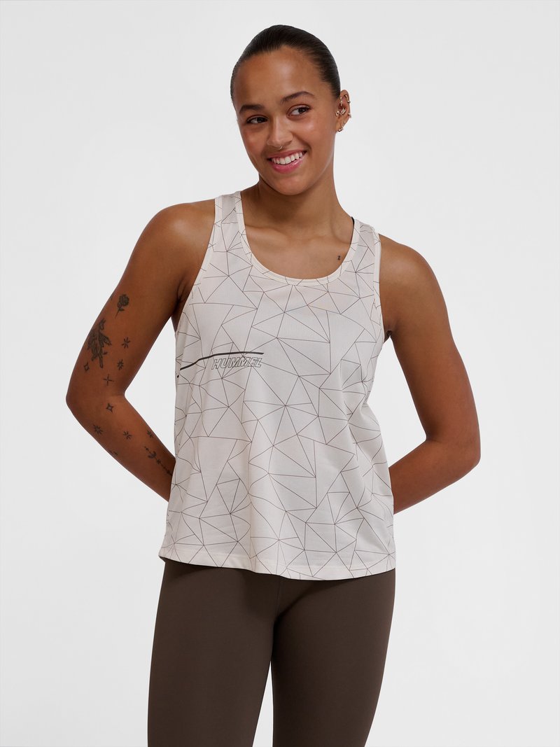 Femme souriante avec des tatouages, portant un haut sans manches blanc à motif géométrique et des leggings foncés, mains dans le dos sur un fond uni.