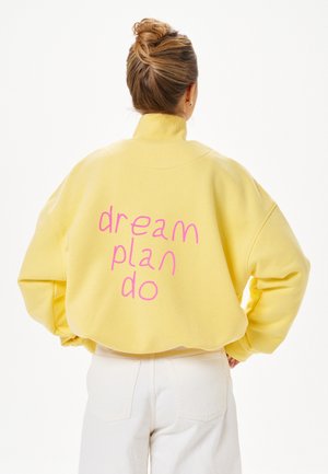 Femme aux cheveux blonds attachés en chignon porte un sweat jaune avec un texte rose « dream plan do » dans le dos, assorti à un pantalon blanc.