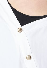 Personne portant un vêtement blanc avec des boutons dorés sur un haut noir, montrant un gros plan du décolleté et des détails des boutons.