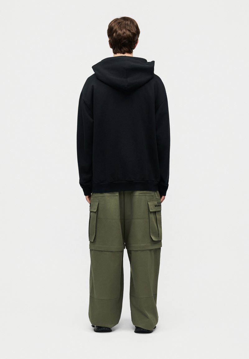 Personne de dos portant un sweat à capuche noir et un pantalon cargo ample vert olive avec de grandes poches latérales devant un fond uni.