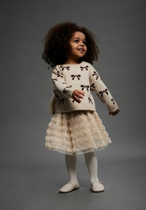 Bēšas adītas džemperis ar tumši brūnu lentu rakstu, slāņota krēmkrāsas tilla svārku ar rozēm aplikācijām, baltas ribotas zeķbikses un gaišas kurpes.