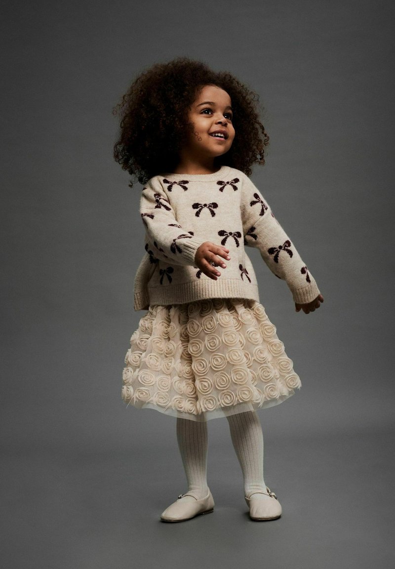 Šviesiai rudas mezginys su tamsiai rudos spalvos kaspino raštu, sluoksniuota grietinėlės tinklelio sijonas su rožių aplikacijomis, baltos rubliai pėdkelnės ir šviesūs batai.