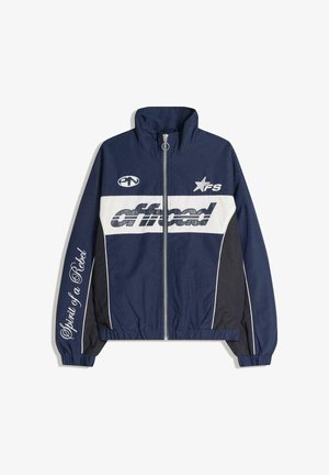 Námořnicky modrá větrovka s bílými akcenty, zapínáním na zip, logovými nášivkami a textem na rukávech. Hladká textura, úzký střih.