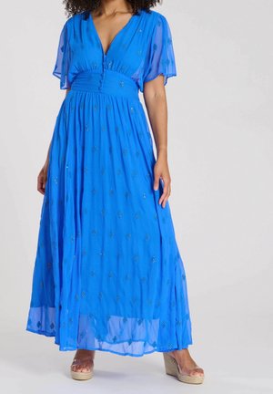 Femme portant une robe bleue longue jusqu'au sol avec des manches courtes évasées, ornée de petits motifs, et des sandales compensées beiges.