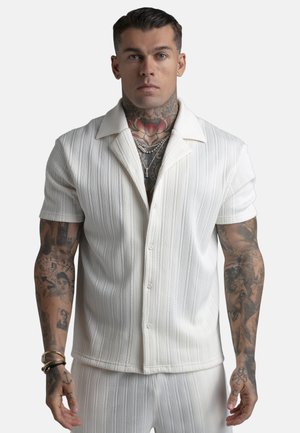 Mees tätoveeringutega, kellel on seljas valge tekstuuriga lühikeste varrukatega nööbitav särk ja sobivad püksid, vaadates otse ette ühevärvilise tausta ees.