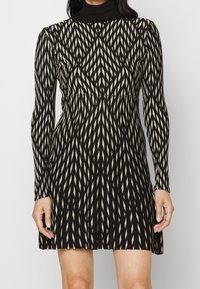 Robe mini à manches longues, noire et blanche, avec un motif géométrique et un col montant, portée par une personne, découvrant les épaules et les jambes au-dessus des genoux.