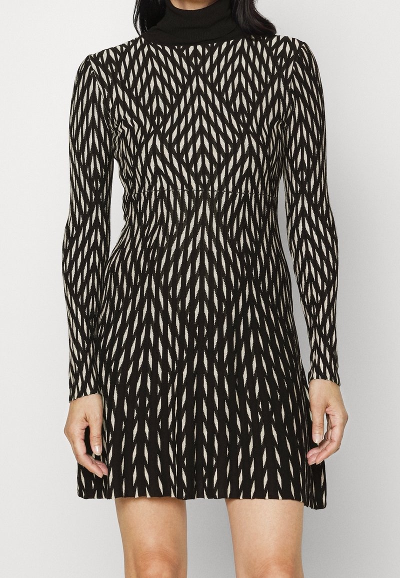 Robe mini à manches longues, noire et blanche, avec un motif géométrique et un col montant, portée par une personne, découvrant les épaules et les jambes au-dessus des genoux.