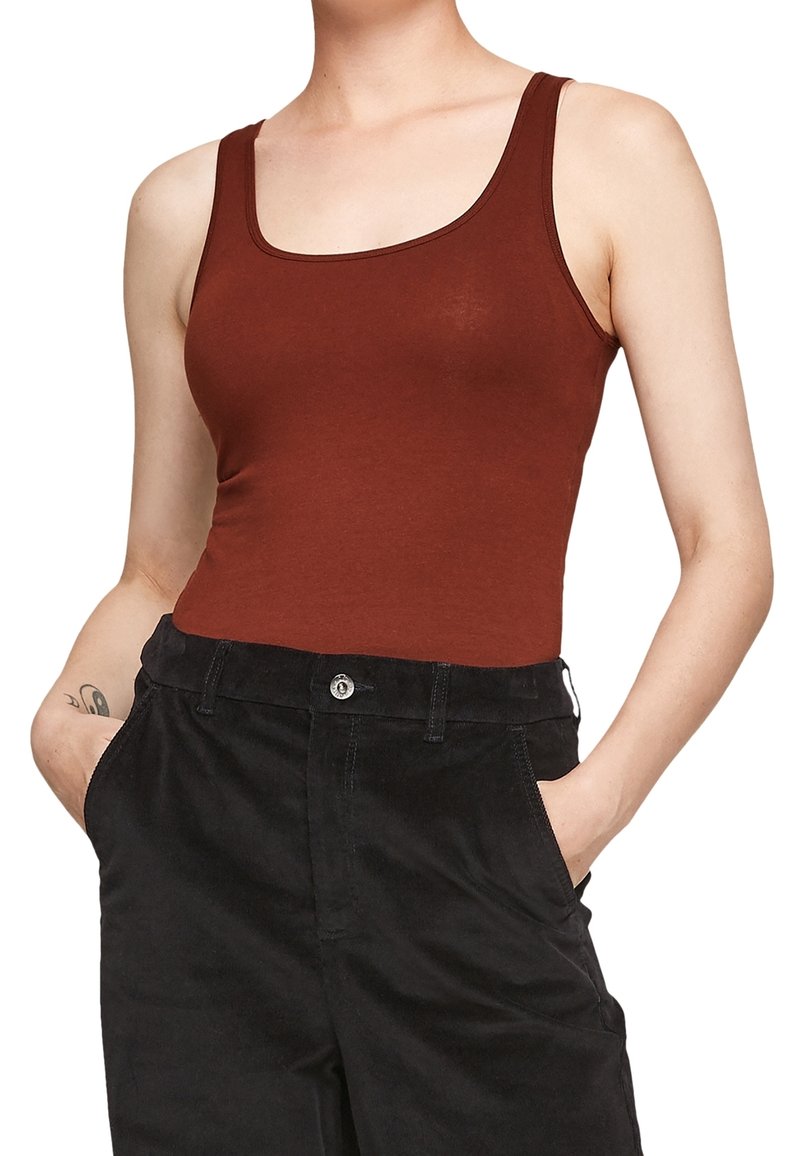 Personne portant un débardeur marron sans manches rentré dans un pantalon noir taille haute, les mains dans les poches et un tatouage sur l'avant-bras gauche.