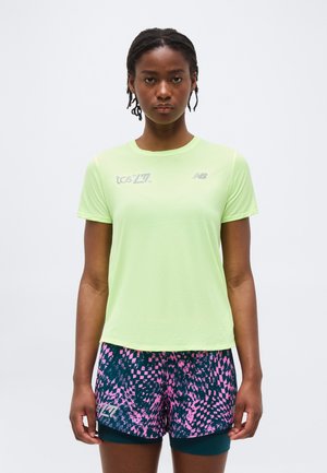 Γυναίκα που φοράει αθλητική μπλούζα κοντομάνικη σε ανοιχτό πράσινο χρώμα και κοντά σορτς για τρέξιμο με σχέδια σε μπλε ναυτικό και ροζ, στέκεται μπροστά σε απλό φόντο.