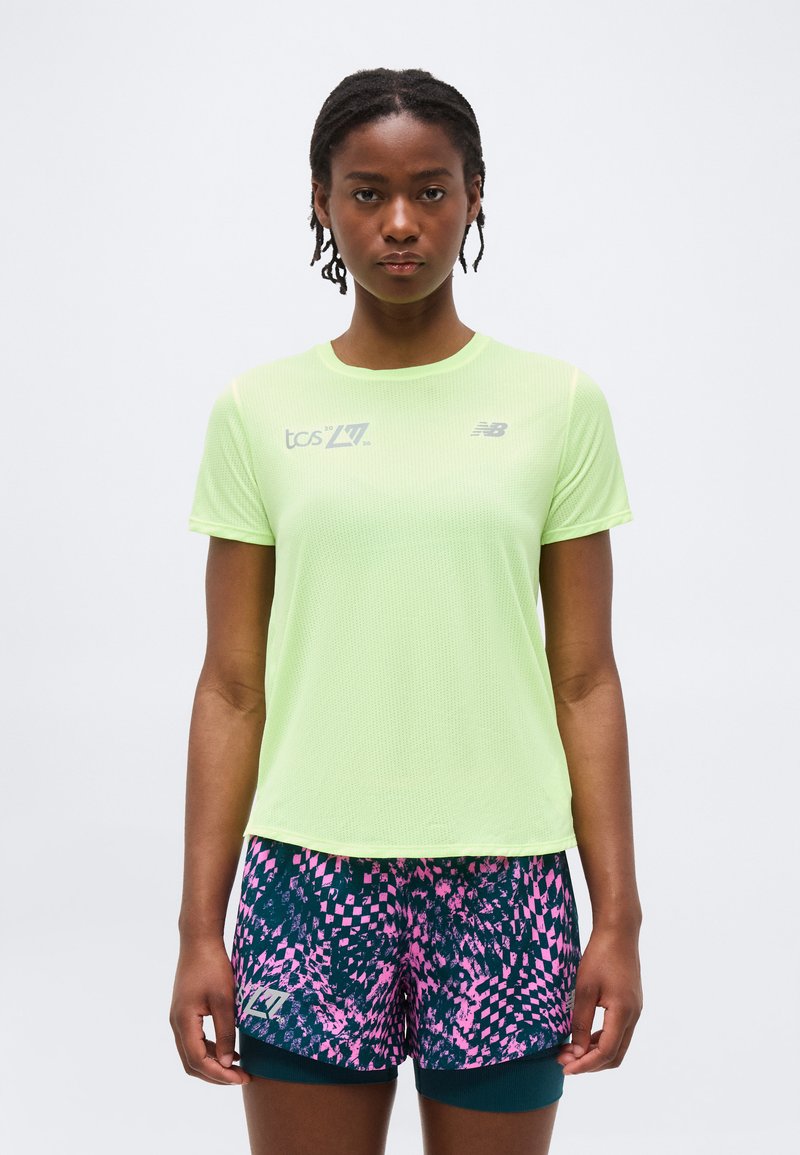 Γυναίκα που φοράει αθλητική μπλούζα κοντομάνικη σε ανοιχτό πράσινο χρώμα και κοντά σορτς για τρέξιμο με σχέδια σε μπλε ναυτικό και ροζ, στέκεται μπροστά σε απλό φόντο.