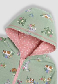 Sudadera verde con capucha que presenta un estampado floral y de animales, con un forro interior de lunares rosas y cierre de cremallera. Material de algodón suave.