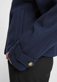 Marine ullblanding jakke mansjett med en foldet design, en gullknapp og en sidelomme for et strukturert utseende.