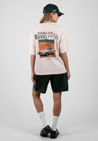 Femme portant une casquette verte, une chemise beige surdimensionnée avec un imprimé voiture et le texte « Thanks for Riding with Us », un short vert foncé, des chaussettes blanches et des chaussures noires.