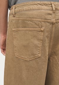 Κοντινό πλάνο ενός ατόμου που φορά ανοιχτό καφέ τζιν παντελόνι, εμφανίζοντας την πίσω τσέπη, τις θηλιές της ζώνης και μέρος ενός γκρι πουλόβερ.