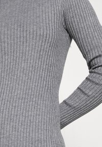 Gros plan d'une personne portant un pull gris côtelé ajusté à manches longues, avec un focus sur le torse et le bras sur un fond uni.