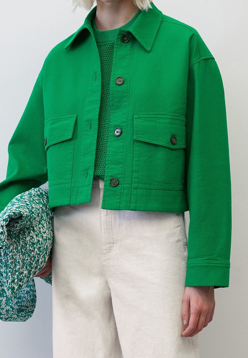 Femme portant une veste courte vert vif par-dessus un haut en tricot, un pantalon beige taille haute, tenant un vêtement tricoté vert et blanc d'une main.